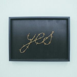 A framed artwork featuring the word 'yes' in barbed wire, displayed in an art gallery indoors.