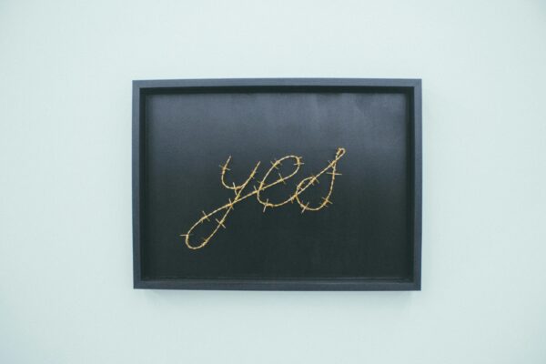 A framed artwork featuring the word 'yes' in barbed wire, displayed in an art gallery indoors.