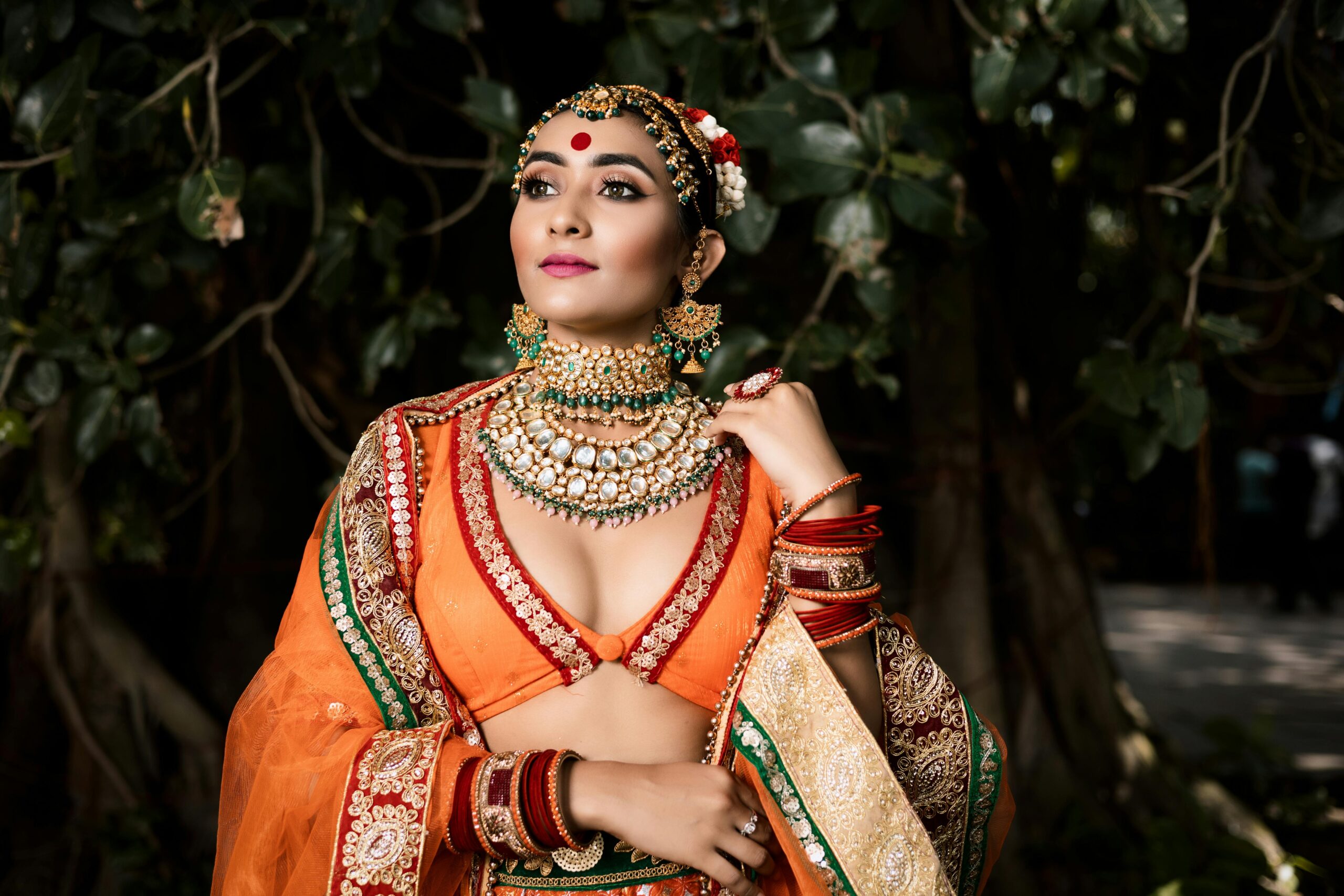 Elegant Indian bride in traditional attire posing outdoors, showcasing exquisite jewelry and vibrant colors.