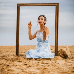 Artistic portrait of a woman on a sandy beach with an empty frame, evoking creativity.